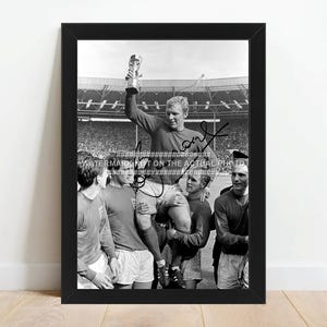 May include: Framed black and white photograph of a football team celebrating. A player holds a trophy above his head, surrounded by teammates. The image is signed and set against a stadium backdrop.