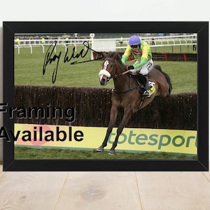 May include: A black and white photograph of a jockey on a brown horse jumping over a fence. The horse is in mid-air and the jockey is leaning forward. The photograph is signed "Roy Whitehead" in black ink. The photograph is framed in a black frame.