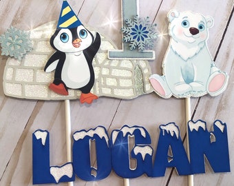 May include: A blue and white penguin cake topper with a party hat, a white polar bear cake topper, a silver number one cake topper, and a blue cake topper with the name "LOGAN" in white letters.