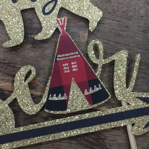 May include: Gold glitter cake topper with the words "baby bear" and a red and black teepee. The teepee has a white fringe and a black and white pattern.