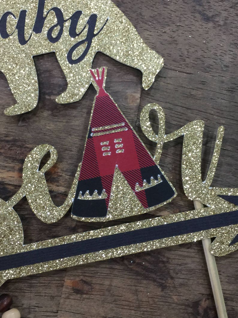 May include: Gold glitter cake topper with the words "baby bear" and a red and black teepee. The teepee has a white fringe and a black and white pattern.