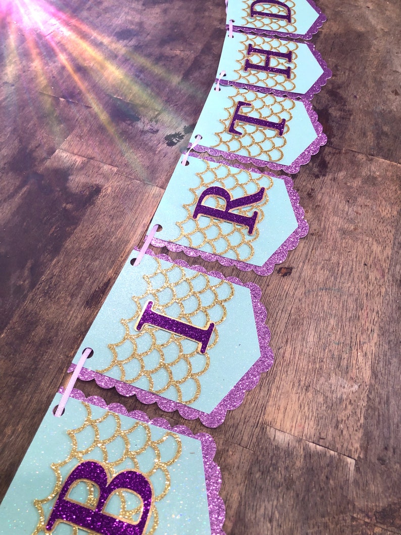 Puede incluir: Una pancarta de cumplea&ntilde;os azul claro y morada con escamas doradas brillantes y la palabra "BIRTHDAY" en letras moradas.