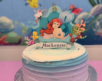 May include: A blue and purple ombre cake with a mermaid themed topper. The topper features Ariel from The Little Mermaid, a seagull, a crab, a starfish, and a fish. The cake is decorated with the name "Mackenzie" and the number "6".
