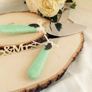 以下が含まれることがあります： ミントグリーンの紐で包まれたハンドルが付いた、ウェディングケーキサーバーとナイフのセット。ハンドルには金属製のチャームと白いリボンが付いています。セットは「Mr & Mrs」の文字が入った木の輪切りに置かれています。