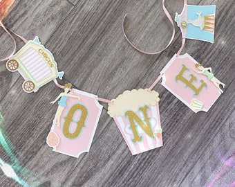 May include: Pink and gold circus themed banner with the word "ONE" in large letters. The banner also includes a small circus wagon, a popcorn bucket, and a ballerina.