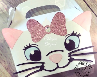 May include: White cardboard party favor box shaped like a cat's face with pink ears, a pink glitter bow with a rhinestone, blue eyes, and black whiskers. The box has a handle and says "Chikpea Original" on the front.