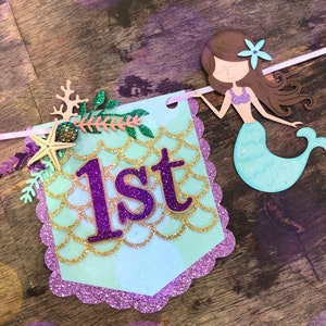Puede incluir: Una pancarta azul claro y morada con un dise&ntilde;o de sirena y la palabra "1er" en letras de purpurina morada. La pancarta est&aacute; decorada con escamas de purpurina dorada, una estrella de mar y una concha.