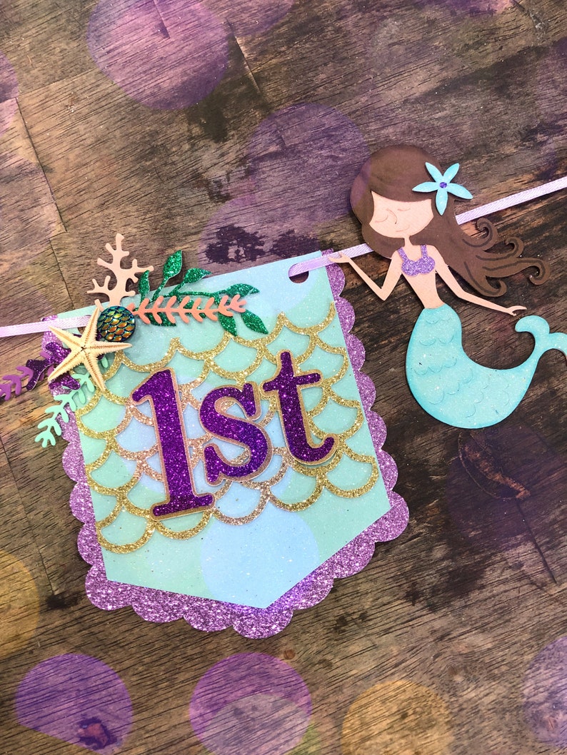 Puede incluir: Una pancarta azul claro y morada con un dise&ntilde;o de sirena y la palabra "1er" en letras de purpurina morada. La pancarta est&aacute; decorada con escamas de purpurina dorada, una estrella de mar y una concha.