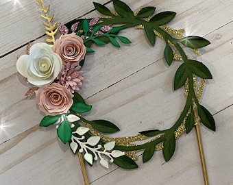 Könnte beinhalten: Ein grüner Papierkranz mit goldenen Glitzerakzenten und rosa und weißen Papierblumen. Der Kranz ist an einem goldenen Kuchenaufsatz befestigt.