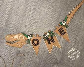 Puede incluir: Una pancarta de esqueleto de dinosaurio marrón con hojas verdes y flores blancas. La pancarta dice "ONE" en letras negras.