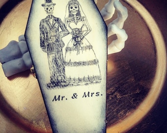 May include: A white coffin-shaped box with a black trim. The box has a black and white illustration of a skeleton bride and groom with the text "Mr. & Mrs."