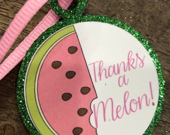 May include: A round, glittery watermelon-themed ornament with a pink ribbon. The ornament features a pink watermelon slice with brown seeds and the text "Thanks a Melon!" in pink script. The border is green.