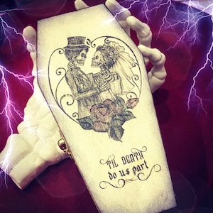 May include: A white wooden coffin-shaped box with a black and white illustration of two skeletons getting married. The text "Til Death Do Us Part" is written in a decorative font on the box.
