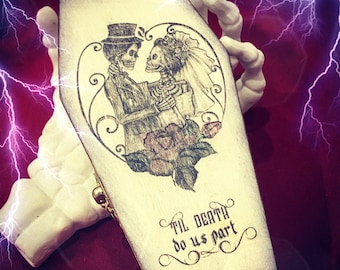 May include: A white wooden coffin-shaped box with a black and white illustration of two skeletons getting married. The text "Til Death Do Us Part" is written in a decorative font on the box.