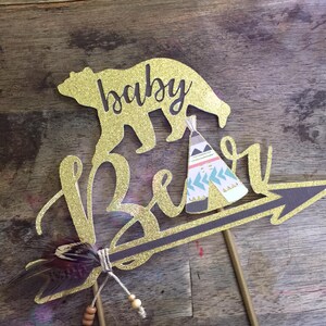 May include: Gold glitter cake topper with the words "baby bear" and a bear silhouette. The bear is holding a small teepee with a pink, blue, and white pattern. An arrow with a feather and beads is attached to the topper.