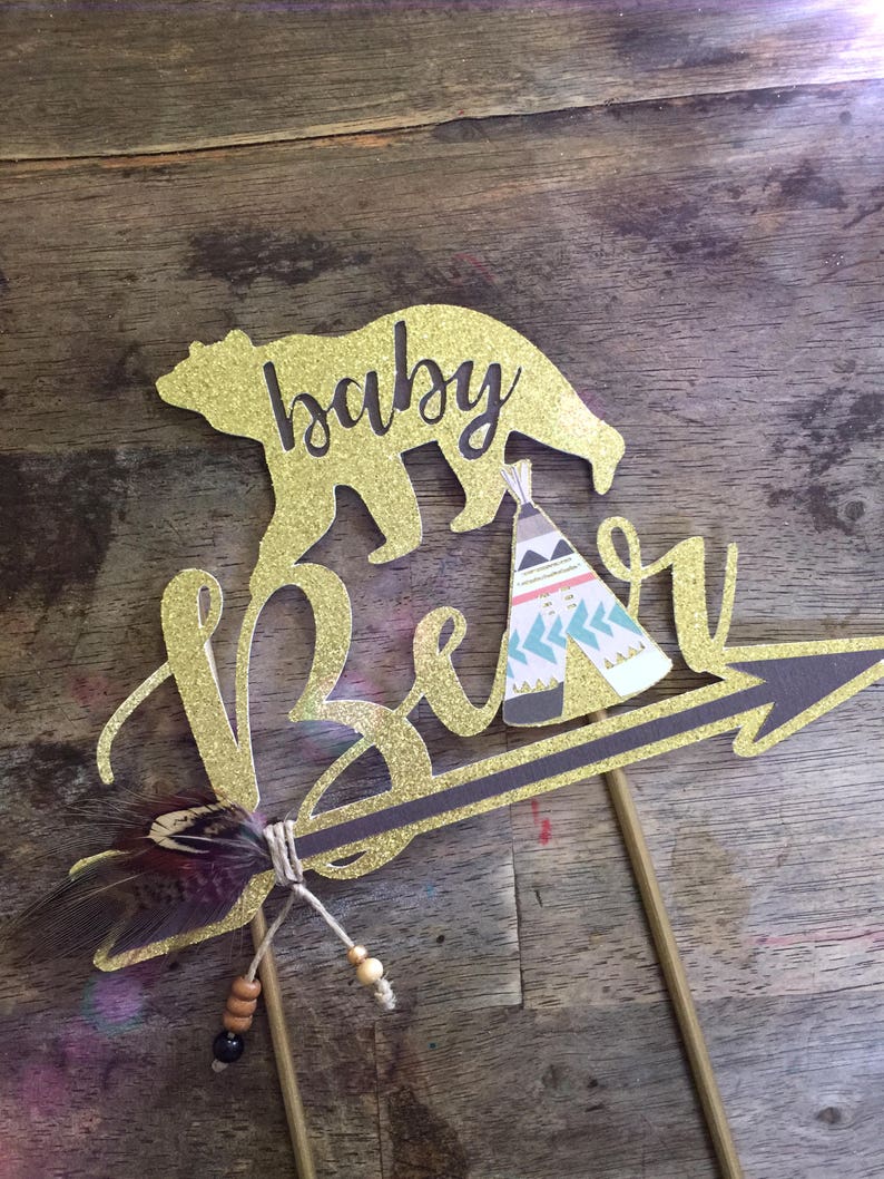 May include: Gold glitter cake topper with the words "baby bear" and a bear silhouette. The bear is holding a small teepee with a pink, blue, and white pattern. An arrow with a feather and beads is attached to the topper.
