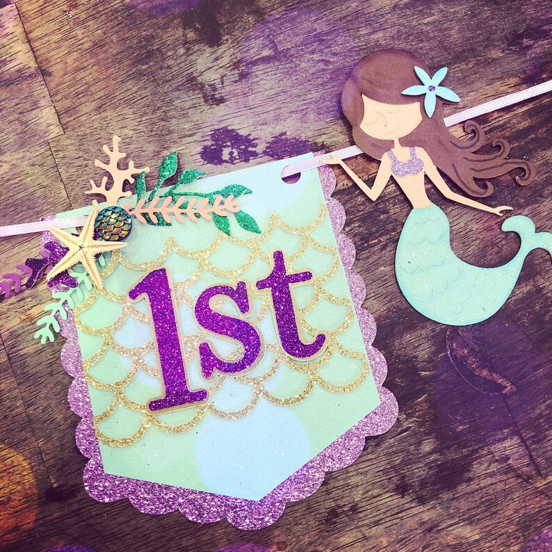 Puede incluir: Una pancarta verde y morada con tem&aacute;tica de sirena con la palabra "1er" en letras de purpurina morada. La pancarta presenta una sirena con cabello casta&ntilde;o y cola verde, una estrella de mar y coral.