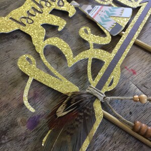 May include: Gold glitter cake topper with the words "baby boy" and a small teepee design. The topper also includes a brown feather and beads.