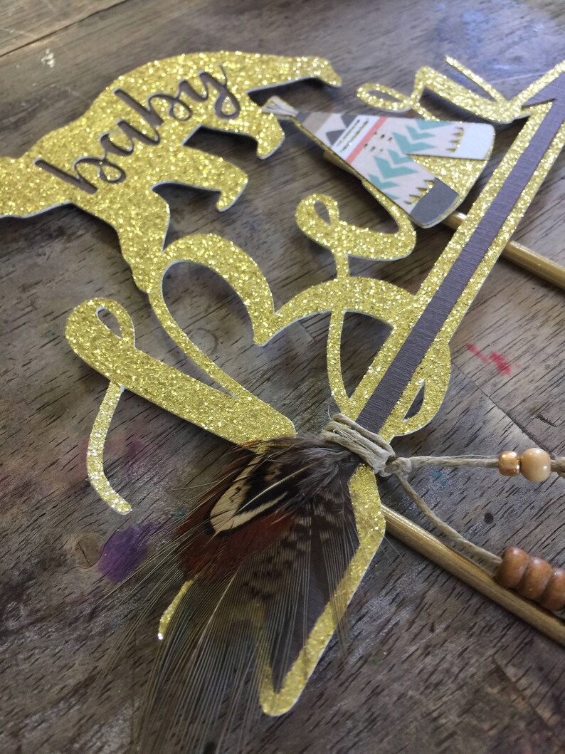 May include: Gold glitter cake topper with the words "baby boy" and a small teepee design. The topper also includes a brown feather and beads.