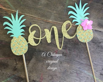 May include: Gold glitter "one" cake topper with two pineapple designs. The pineapples are green and gold with a pink flower on one. The text "A Chikapeoa original design" is below the topper.