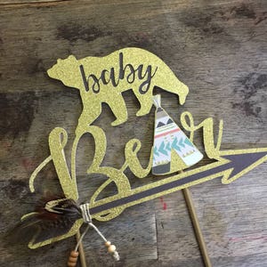 May include: Gold glitter cake topper with the words "baby bear" and a bear silhouette. The bear is holding a small teepee with a geometric pattern. A brown feather and beads are attached to the arrow.