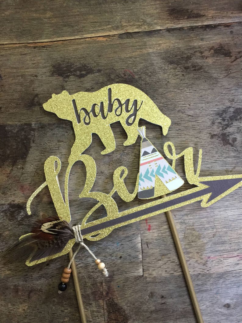 May include: Gold glitter cake topper with the words "baby bear" and a bear silhouette. The bear is holding a small teepee with a geometric pattern. A brown feather and beads are attached to the arrow.