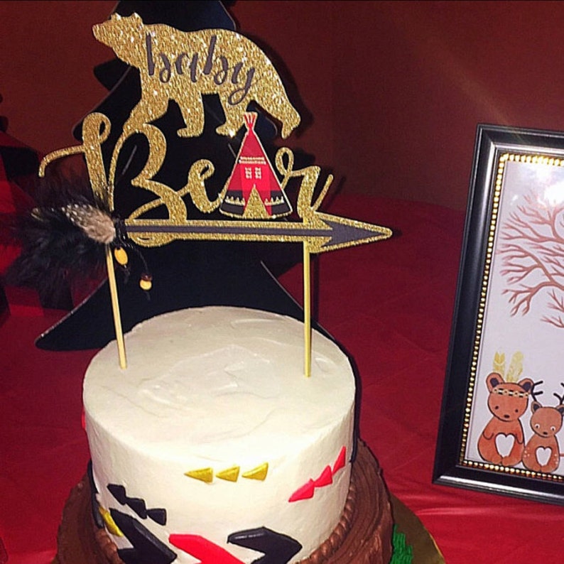 May include: A two-tiered cake decorated with a gold glitter cake topper that says "Baby Bear" with a bear, a teepee, and an arrow. The cake is decorated with black, red, and gold geometric shapes.