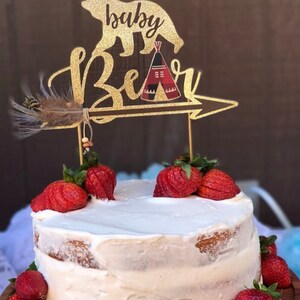 May include: A white cake with a gold glitter cake topper that says "baby bear" with a bear and a teepee design. The cake is decorated with fresh strawberries.