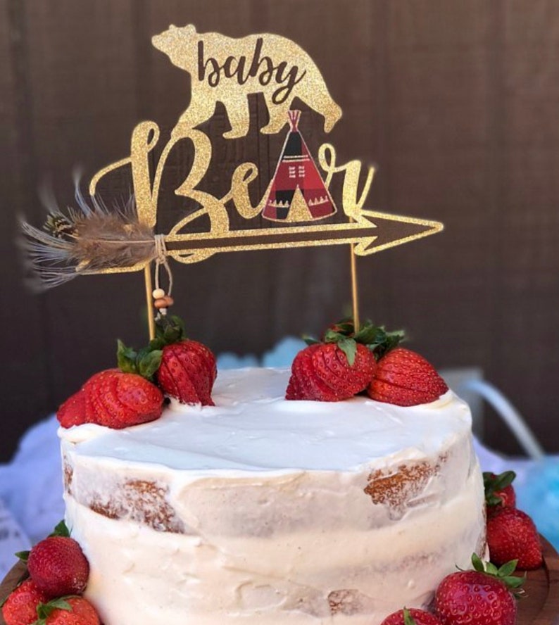 May include: A white cake with a gold glitter cake topper that says "baby bear" with a bear and a teepee design. The cake is decorated with fresh strawberries.