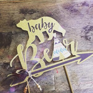 May include: Gold glitter cake topper with a bear silhouette, a teepee, and the words "baby bear" on an arrow. The arrow has a brown feather attached to it.