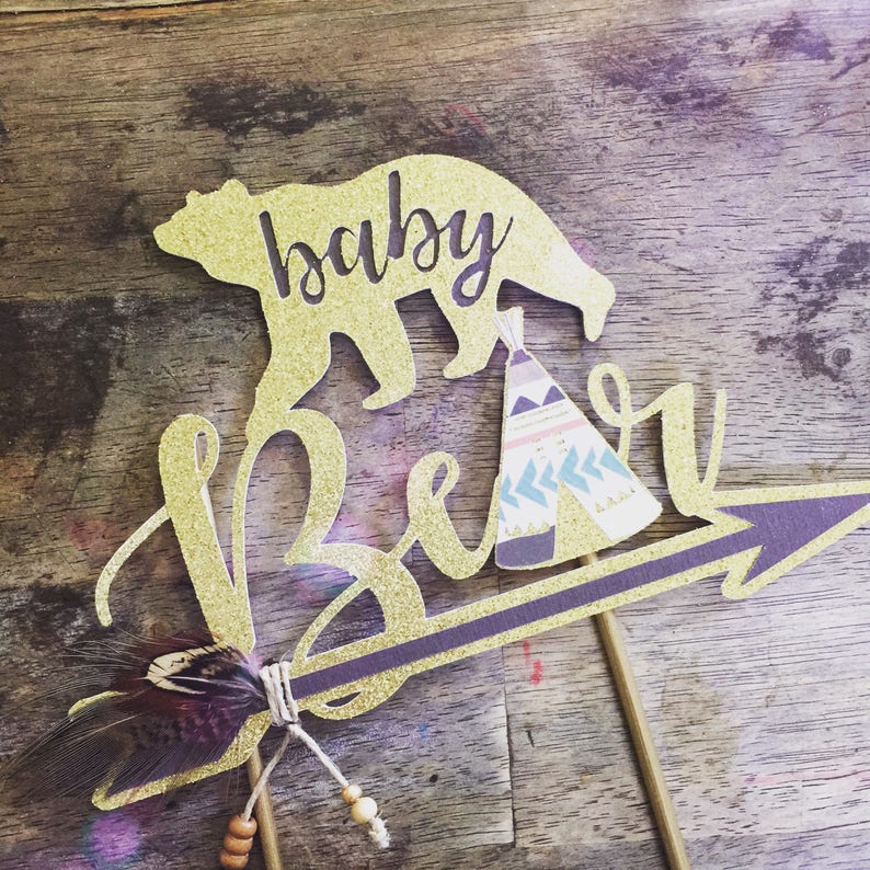 May include: Gold glitter cake topper with a bear silhouette, a teepee, and the words "baby bear" on an arrow. The arrow has a brown feather attached to it.