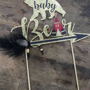 May include: Gold glitter cake topper with a bear silhouette, a teepee, and an arrow with a feather. The text "baby bear" is written on the topper.