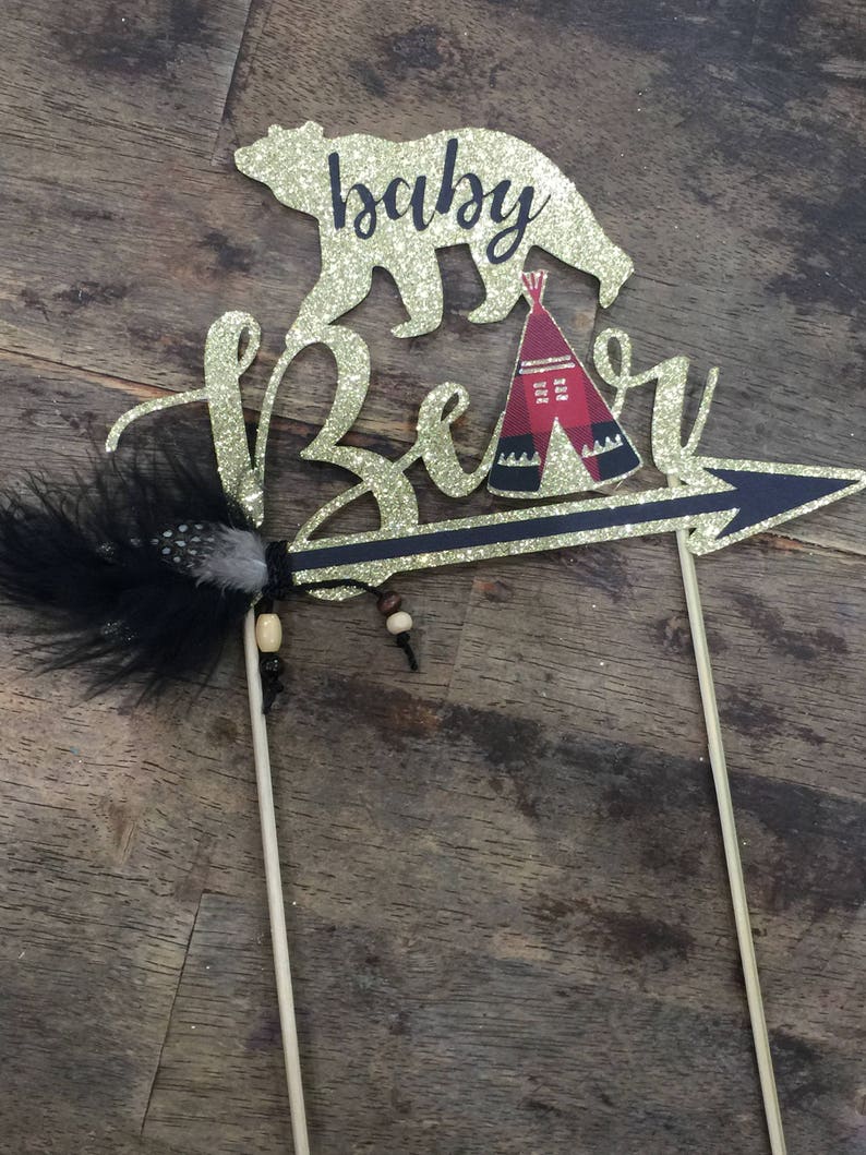 May include: Gold glitter cake topper with a bear silhouette, a teepee, and an arrow with a feather. The text "baby bear" is written on the topper.