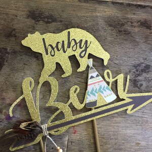 May include: Gold glitter cake topper with the words "baby bear" and a small teepee design. The topper features a gold glitter arrow with a brown feather and beads.