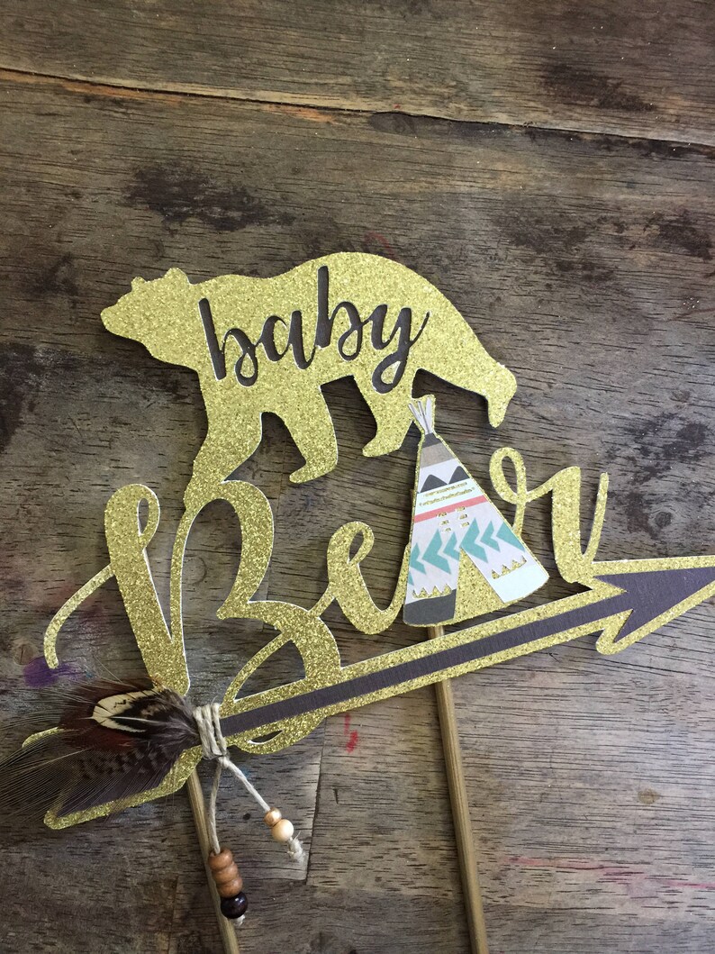 May include: Gold glitter cake topper with the words "baby bear" and a small teepee design. The topper features a gold glitter arrow with a brown feather and beads.