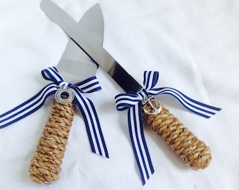 May include: A set of two cake cutting knives with rope handles and blue and white striped ribbon bows. One knife has a silver anchor charm and the other has a silver life preserver charm.