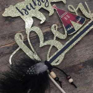 May include: Gold glitter cake topper with the words "baby bear" and a small red and black teepee. A black feather with white dots and brown beads are attached to a wooden stick.