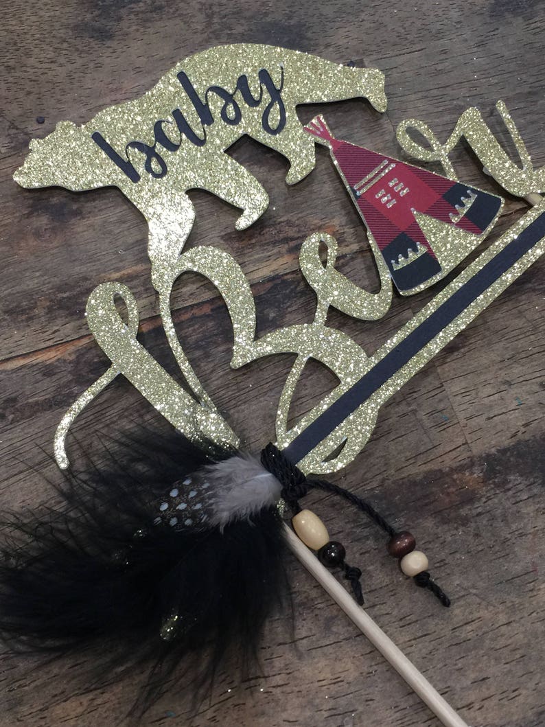May include: Gold glitter cake topper with the words "baby bear" and a small red and black teepee. A black feather with white dots and brown beads are attached to a wooden stick.