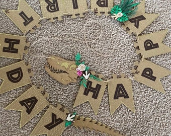 May include: A brown and black banner with the words "Happy Birthday" in a dinosaur theme. The banner is decorated with green leaves and a dinosaur skull.