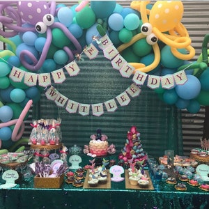 Puede incluir: Una fiesta de primer cumplea&ntilde;os con tem&aacute;tica de sirena con una mesa de postres que incluye cupcakes, cake pops y un pastel de varios niveles. El tel&oacute;n de fondo est&aacute; decorado con globos azules y verdes, decoraciones de pulpo y una pancarta que dice "Happy 1st Birthday Whittle".