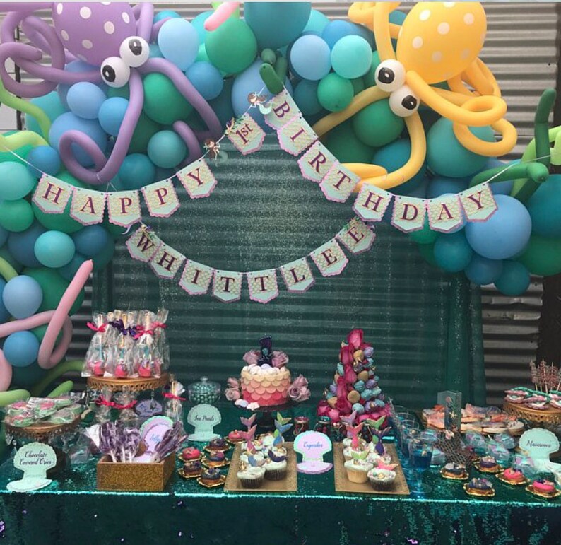 Puede incluir: Una fiesta de primer cumplea&ntilde;os con tem&aacute;tica de sirena con una mesa de postres que incluye cupcakes, cake pops y un pastel de varios niveles. El tel&oacute;n de fondo est&aacute; decorado con globos azules y verdes, decoraciones de pulpo y una pancarta que dice "Happy 1st Birthday Whittle".