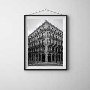 May include: Black and white framed photograph of a large corner building with multiple storeys, balconies, and arched entryways. The building is in a classical style, with ornate details and a symmetrical facade. The photograph is hanging on a white wall.