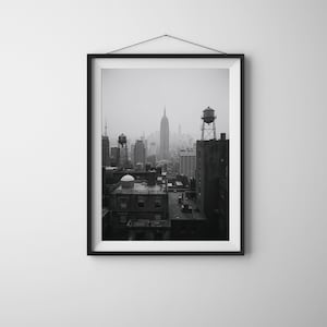 May include: Black and white framed print of a New York City skyline on a cloudy day. The Empire State Building is visible in the center, with water towers on the rooftops of the buildings in the foreground.