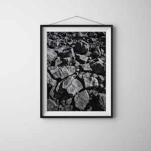 Puede incluir: Impresión enmarcada en blanco y negro que presenta una vista de cerca de un montón de rocas de color gris oscuro. Las rocas varían en tamaño y forma, con grietas y texturas visibles. La obra de arte se exhibe en una pared blanca.