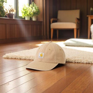 Puede incluir: Gorra de b&eacute;isbol beige con visera curva y un peque&ntilde;o logotipo cuadrado. La gorra est&aacute; hecha de una tela resistente y tiene un dise&ntilde;o cl&aacute;sico de seis paneles. El sombrero est&aacute; sobre un piso de madera.