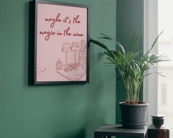 Puede incluir: Un cuadro enmarcado con el texto "maybe it's the magic in the wine" en cursiva. El fondo rosa presenta una ilustración con temática de vino con copas, una botella y uvas. El marco es negro y el cuadro cuelga de una pared verde.