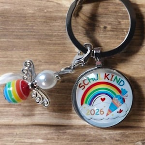 Op de afbeelding: Een zilverkleurige sleutelhanger met een ronde bedel voorzien van een regenboog, een kleine puntzak en de tekst SCHU KIND 2026. Aan de ring hangt een kleine bijvormige bedel met een regenbooggestreept lijfje, een parelhoofd en zilveren vleugels.