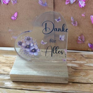 Op de afbeelding: Een hartvormig, transparant acrylbordje met een boeket paarse bloemen en kleine vlinders. De tekst "Danke für Alles" is in donkerblauw cursief schrift gedrukt. Het bordje is bevestigd op een rechthoekige, lichtgekleurde houten voet.
