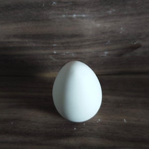 May include: A single, white egg with a smooth surface. The egg is oval-shaped and stands upright on a dark brown wooden surface. The lighting is soft, highlighting the egg's form.