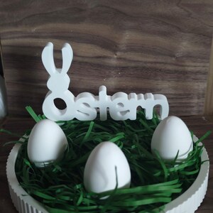 May include: A white Easter centrepiece featuring three white eggs in green paper grass. A white bunny silhouette and the word "Ostern" are above the eggs. The centrepiece is in a white, fluted-edge dish.